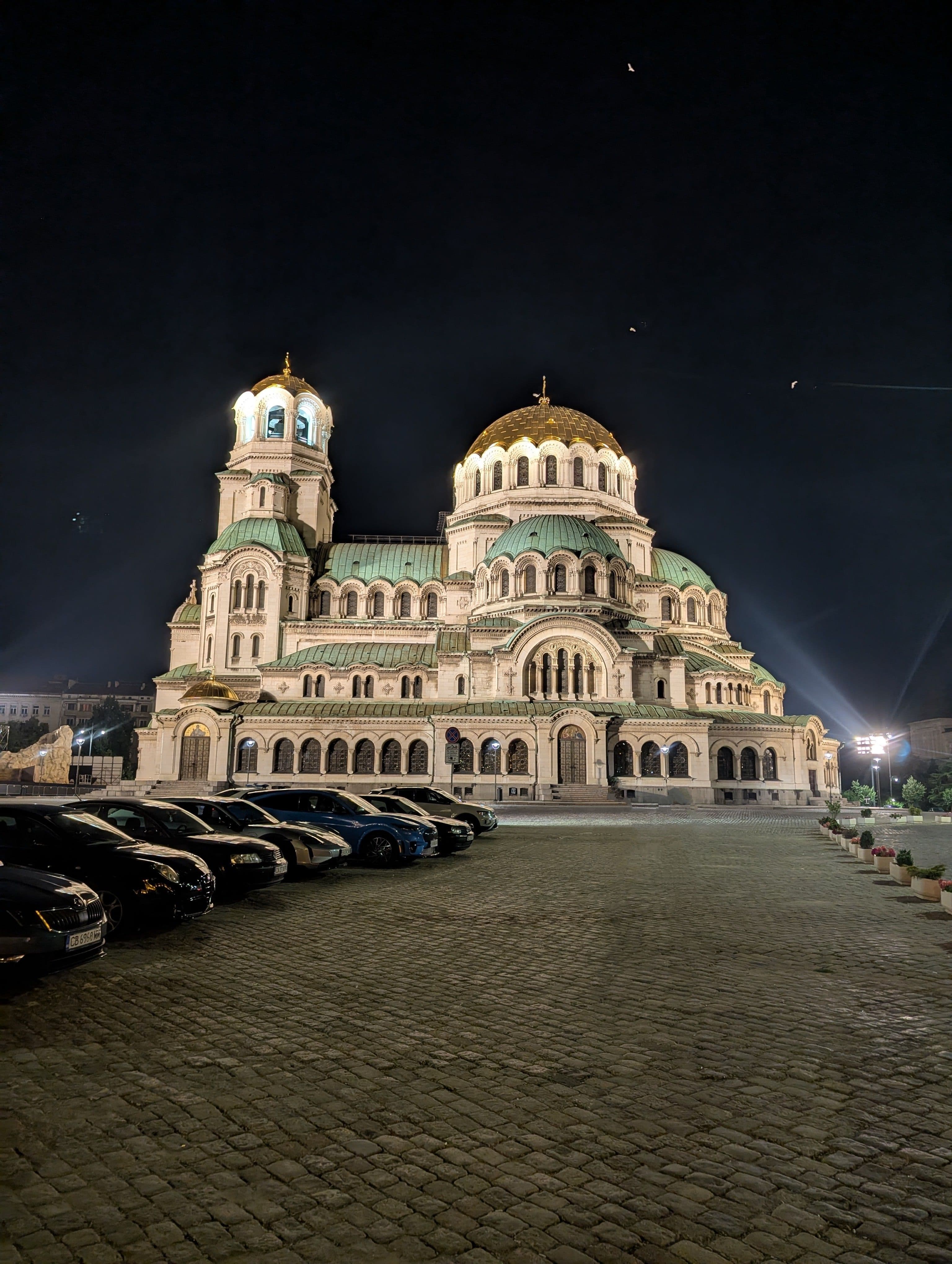 A large Orthodox cathedral with distinctive Byzantine architecture photographed at night in Bulgaria, featuring a central golden dome and bell tower with green copper roofing. The cream-colored stone building is illuminated by exterior lighting against a dark sky. Multiple cars are parked in front of the cathedral on a cobblestone plaza or square.
